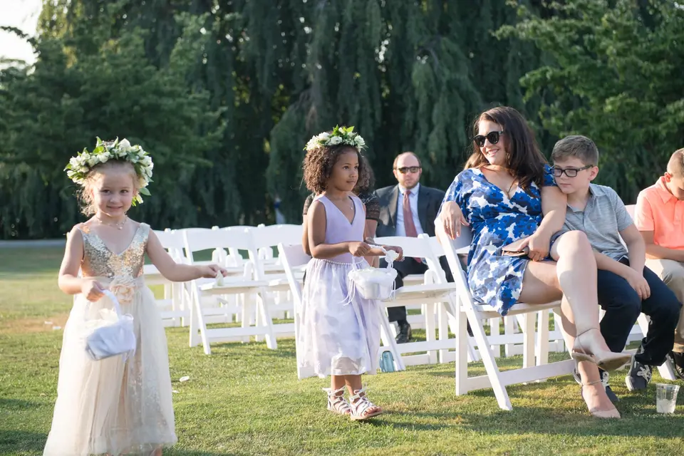 Flower girls in floral crowns walk past guests seated on the lawn.