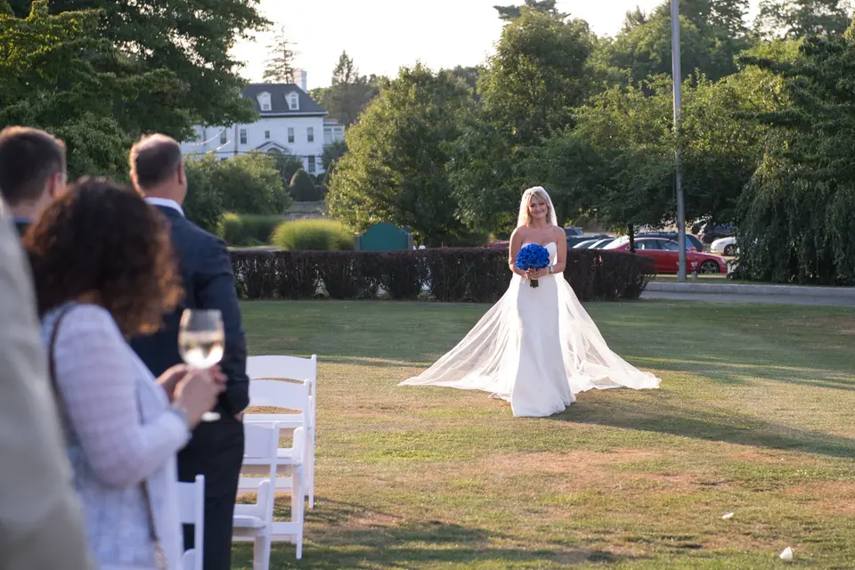 The bride walks alone across the lawn toward the ceremony, her bouquet of blue roses in hand.