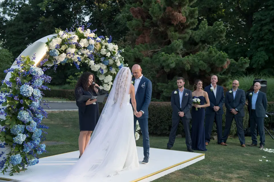 The bride and groom face each other beneath the flower arch as the officiant speaks, the groomsmen standing by.