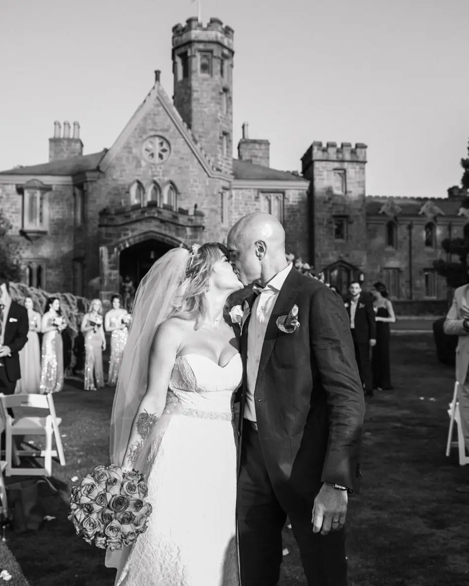 The bride and groom kiss in the aisle as guests cheer, the castle behind. Black and white.
