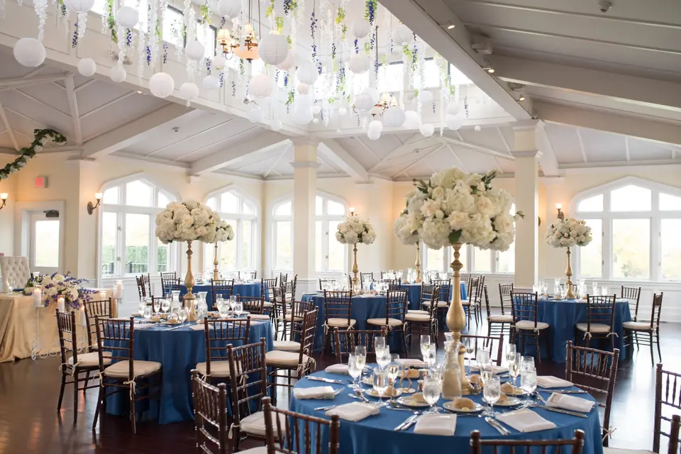 Round tables in deep blue linen with tall white arrangements on gold stands, arched windows beyond.