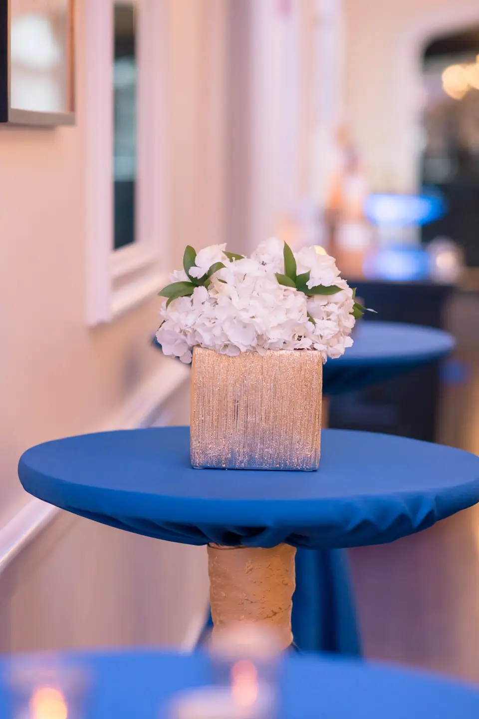A small gold vase of white hydrangeas on a cocktail table in blue linen.