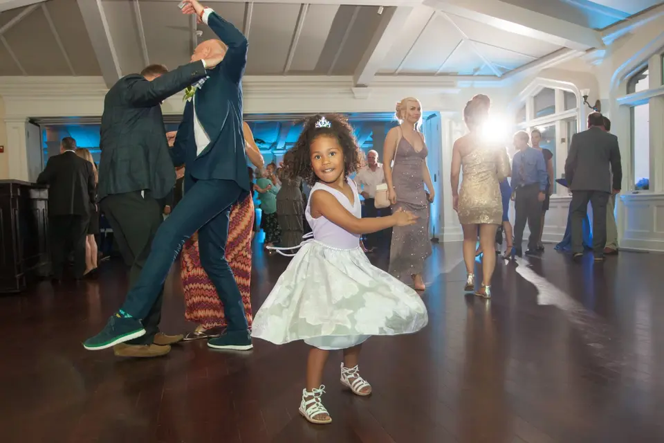 A little girl in a tiara twirls on the dance floor as guests dance behind her.