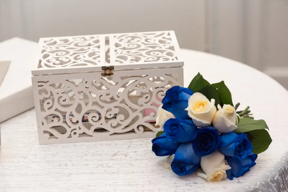 A white filigree card box beside a bouquet of blue and ivory roses.