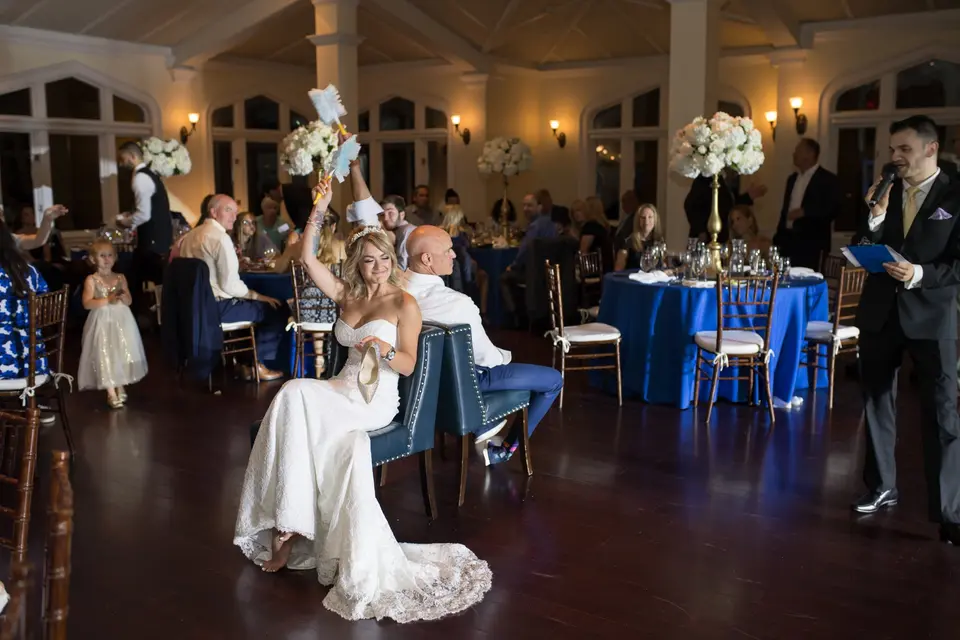 The bride sits on a chair on the dance floor and waves a garter above her head as guests laugh.