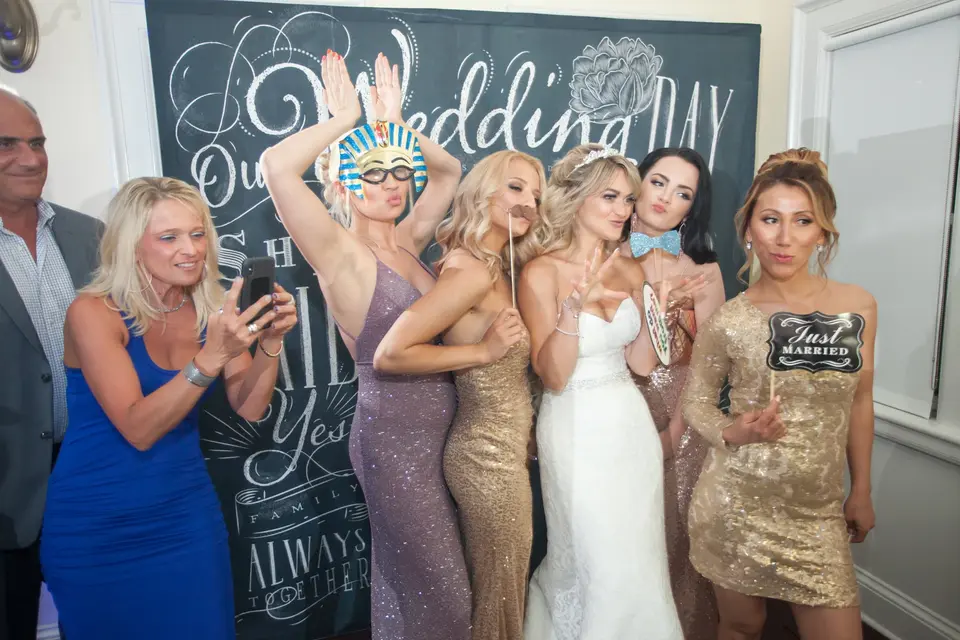 The bride and her friends pose with photo props before a chalkboard wedding sign.