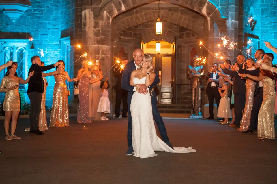The bride and groom embrace between two lines of guests holding sparklers, the castle doorway lit in blue.