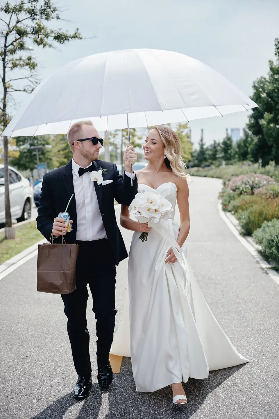 The couple walk along a waterfront path beneath a white umbrella, the bride holding white orchids.