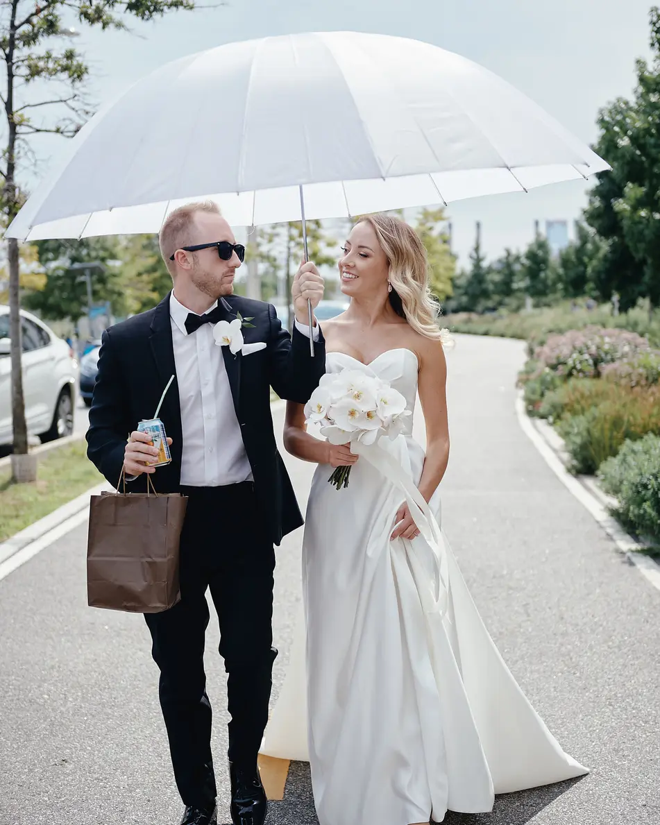 The couple walk along a waterfront path beneath a white umbrella, the bride holding white orchids.
