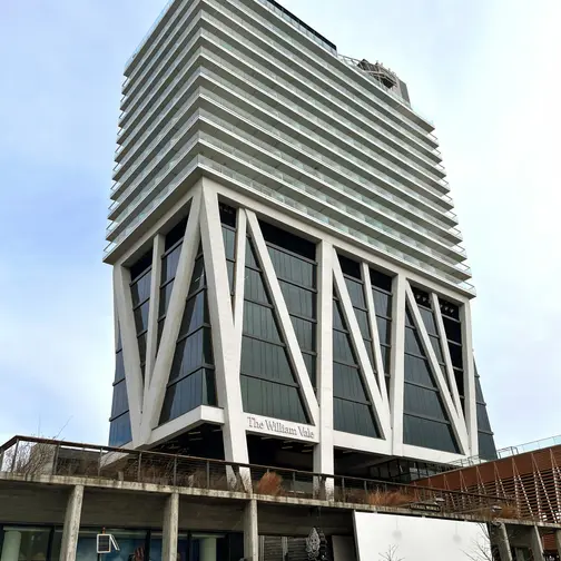 The William Vale in Williamsburg, a white tower raised on angled concrete legs above a public park, against a bright sky.