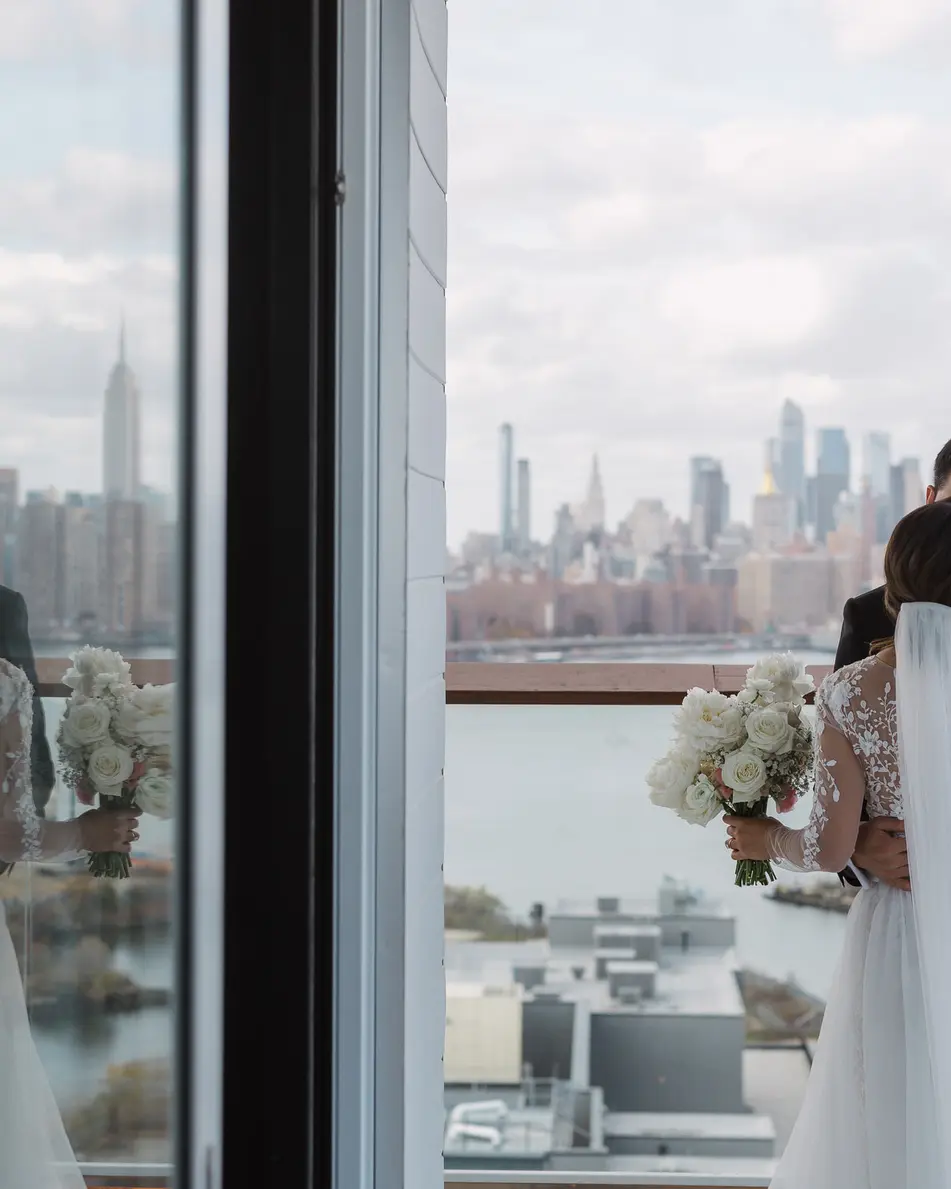 The bride and groom stand at a floor-to-ceiling window, the Empire State Building on the skyline beyond.