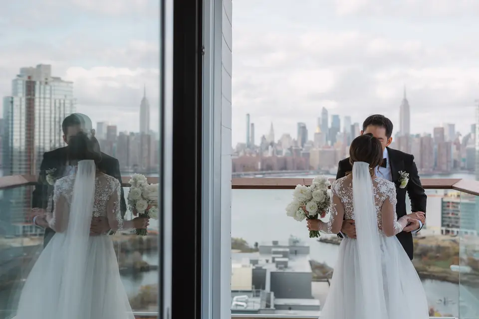 The bride and groom stand at a floor-to-ceiling window, the Empire State Building on the skyline beyond.