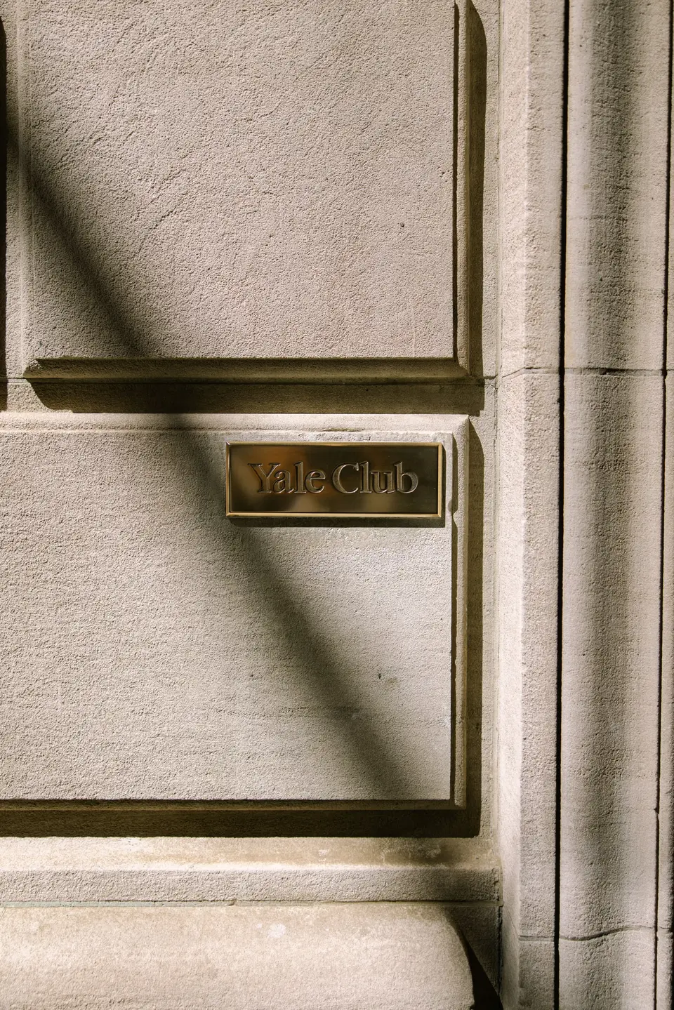 A brass Yale Club plaque on the building's stone facade, crossed by a shadow.
