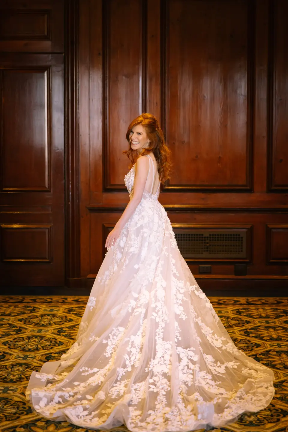 The bride looks back over her shoulder in a wood-panelled room, her lace train spread across the carpet.