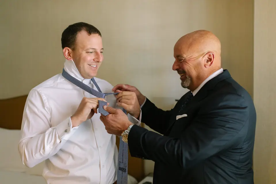 The groom's father helps him with his tie, both laughing.