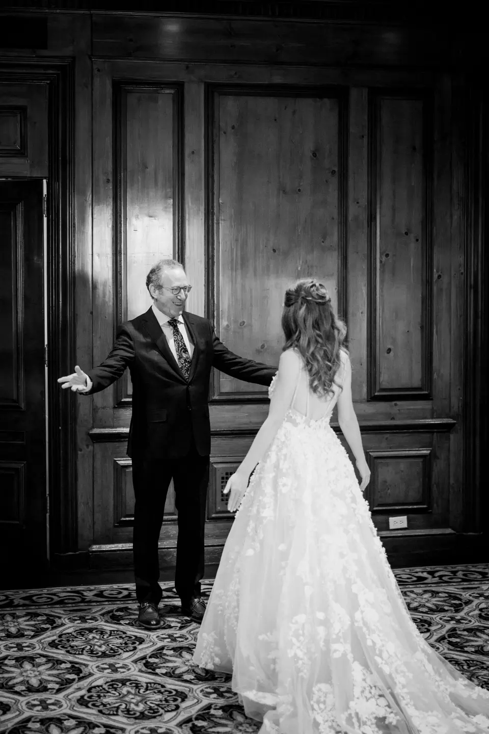 The bride's father opens his arms as he sees her for the first time in her gown. Black and white.