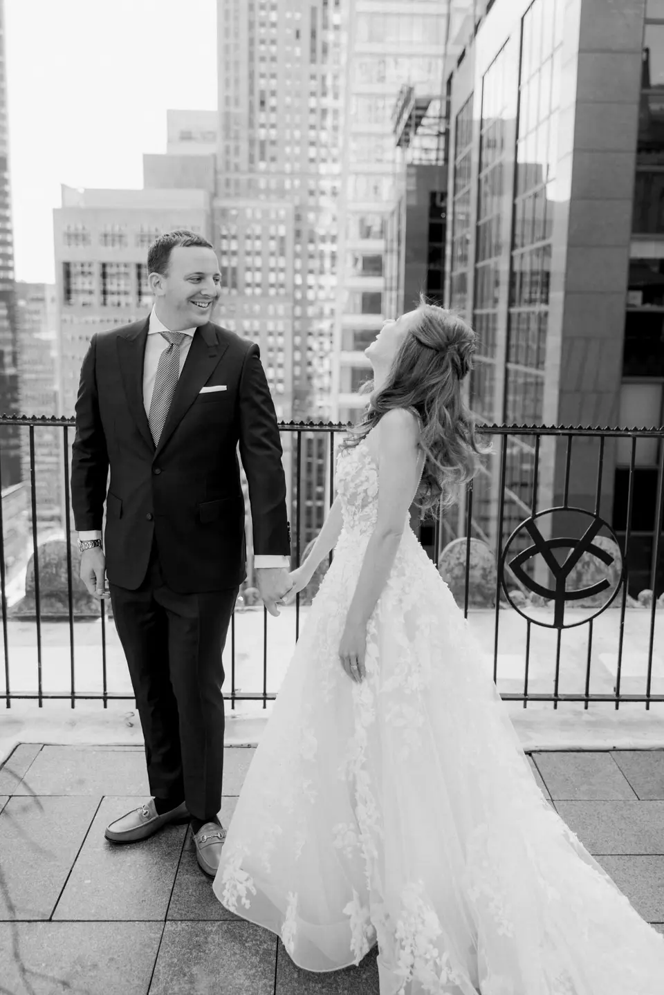 On the roof terrace, the groom holds the bride's hand as she laughs, Midtown towers around them. Black and white.