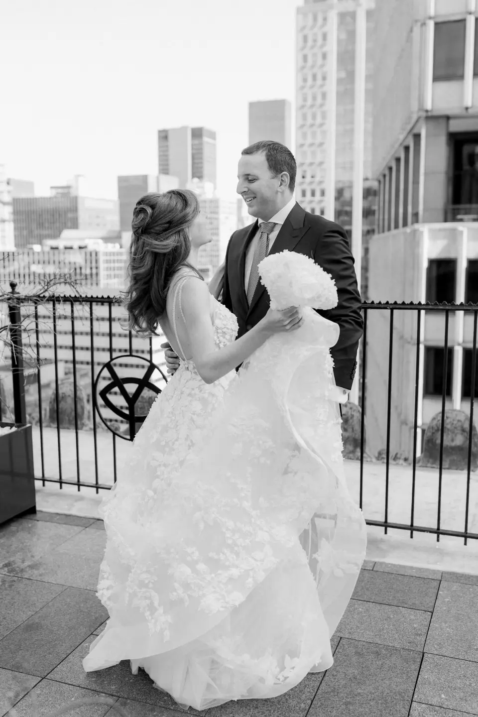 The groom smiles as the bride turns toward him on the terrace, her skirt in her hands. Black and white.