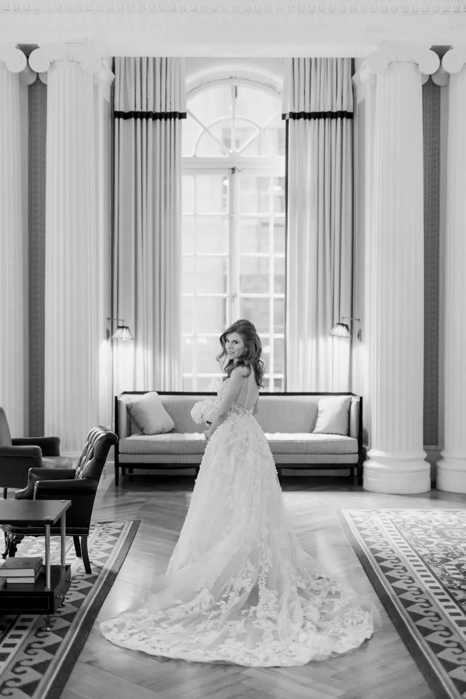 The bride stands between tall columns in a lounge with a Palladian window. Black and white.