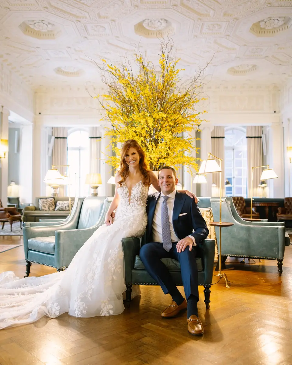 The bride and groom sit together on a pale sofa in a sunlit lounge at The Yale Club, her gown spread across the parquet floor.