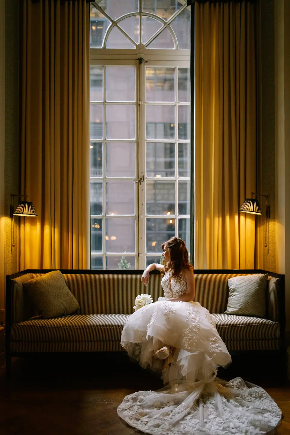 The bride sits on a sofa beneath a tall window, her gown spread across the floor.