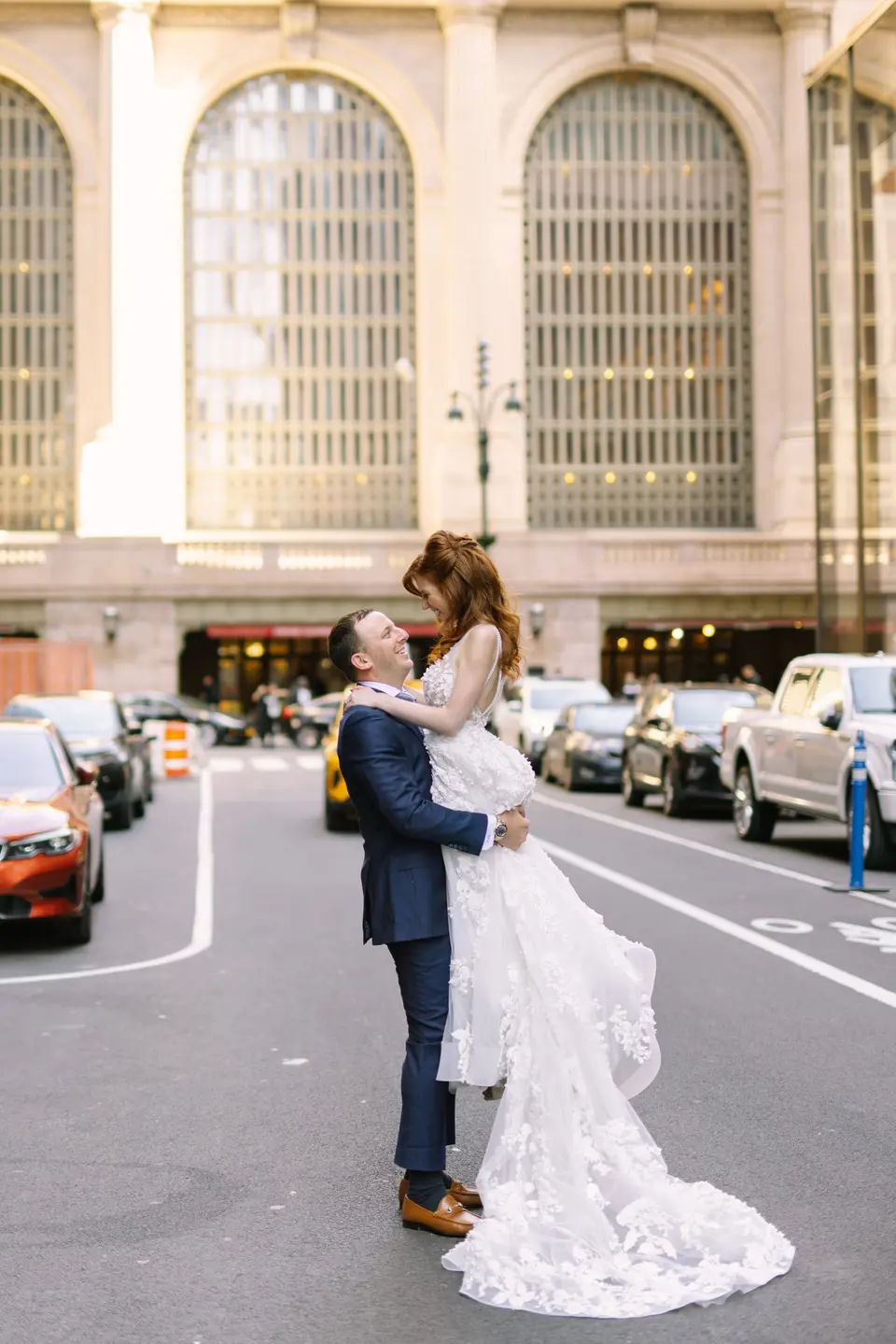 The groom lifts the bride in the middle of Vanderbilt Avenue, Grand Central's arched windows behind them.