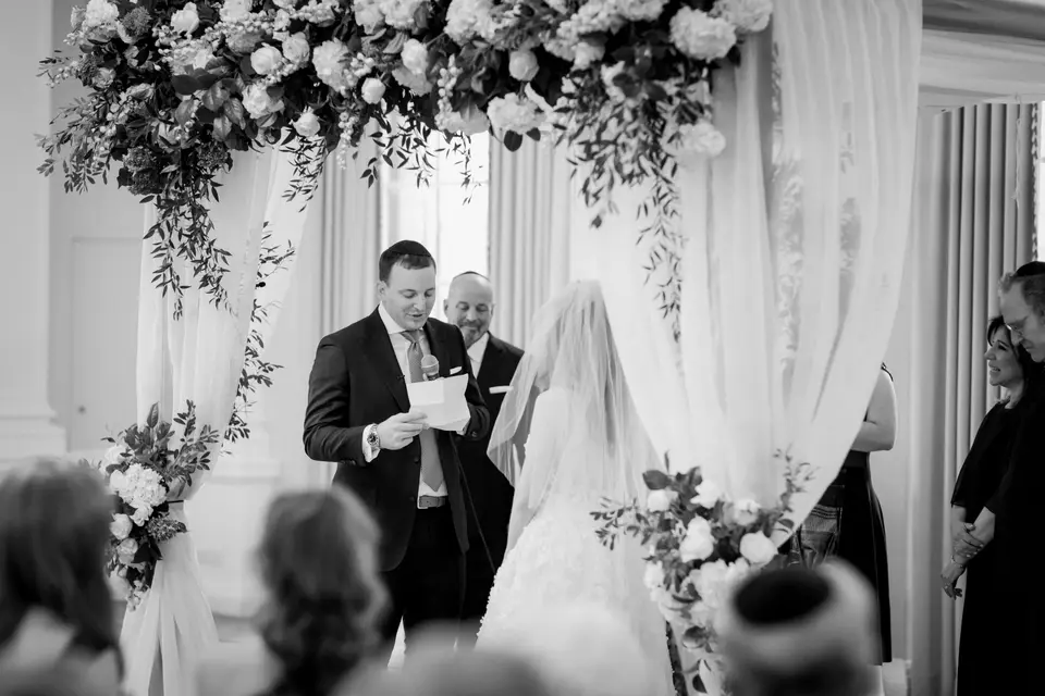 The groom reads his vows to the bride beneath the flowered chuppah. Black and white.