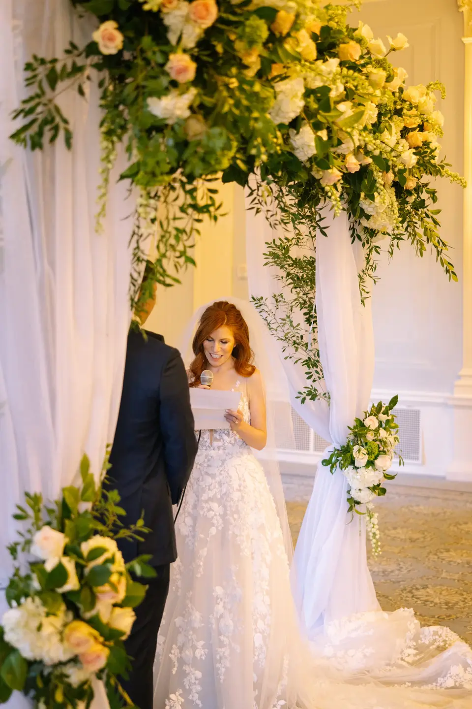 The bride smiles as she reads her vows beneath the chuppah of white flowers and drapes.