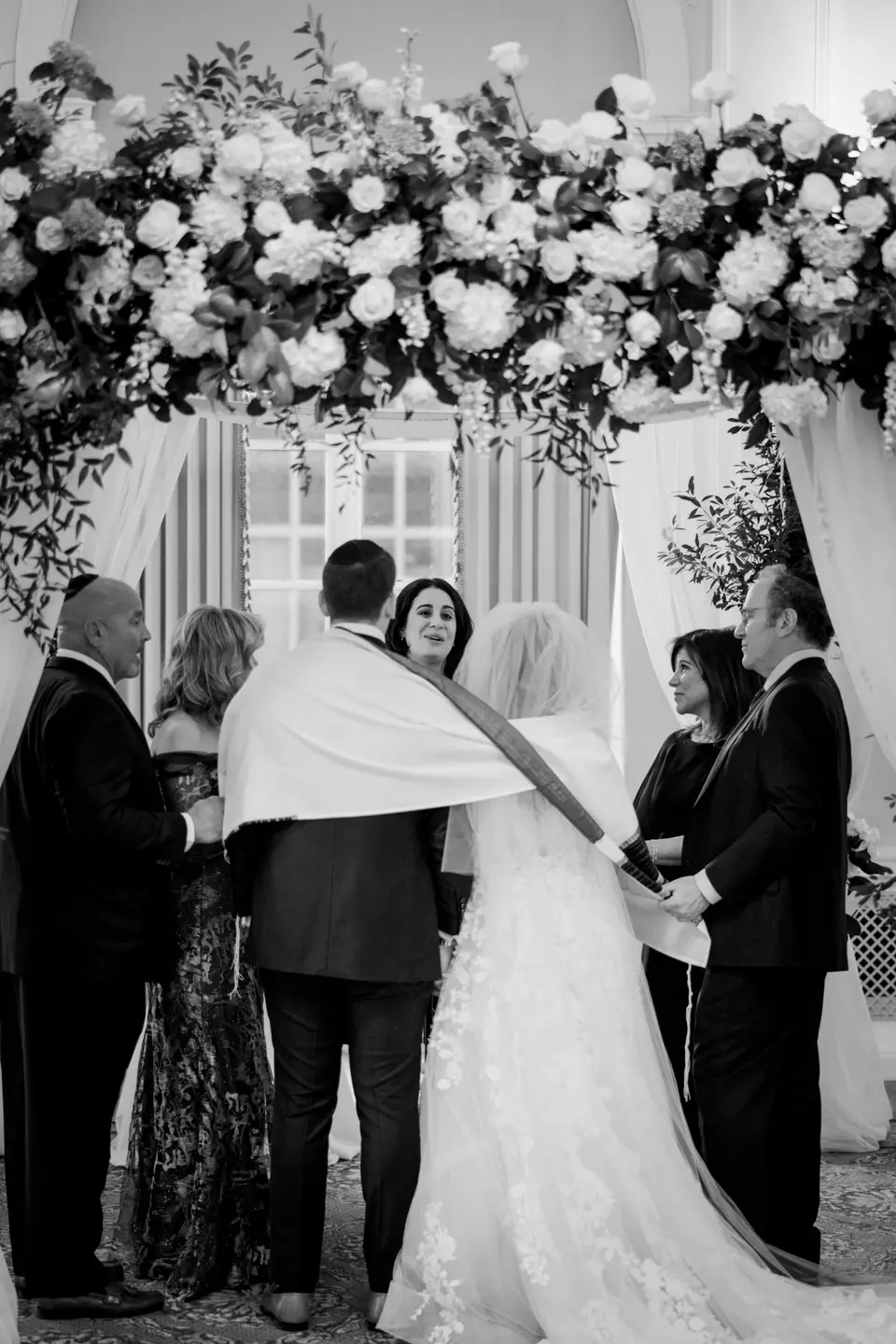 Beneath the chuppah, a prayer shawl is wrapped around the bride and groom, family close by. Black and white.