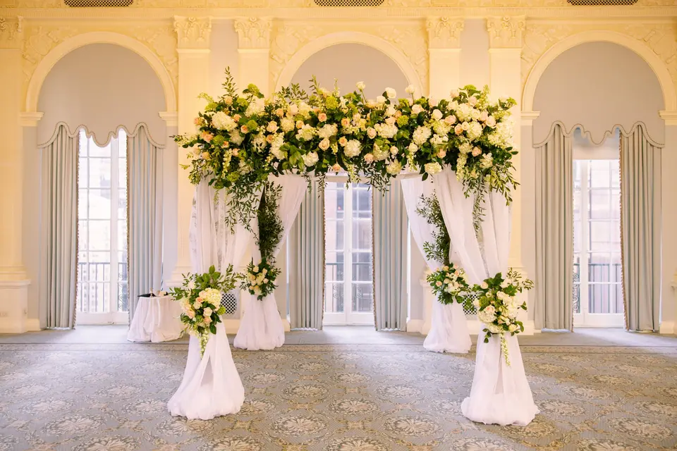 The chuppah of white roses and greenery hung with white drapes, before arched windows.