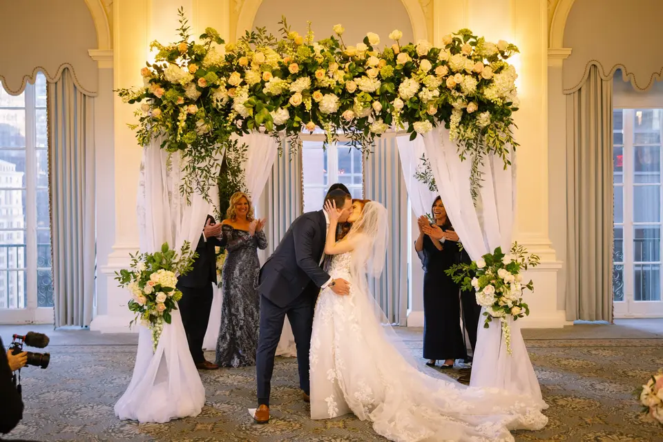 The bride and groom kiss beneath the flowered chuppah as their families look on.