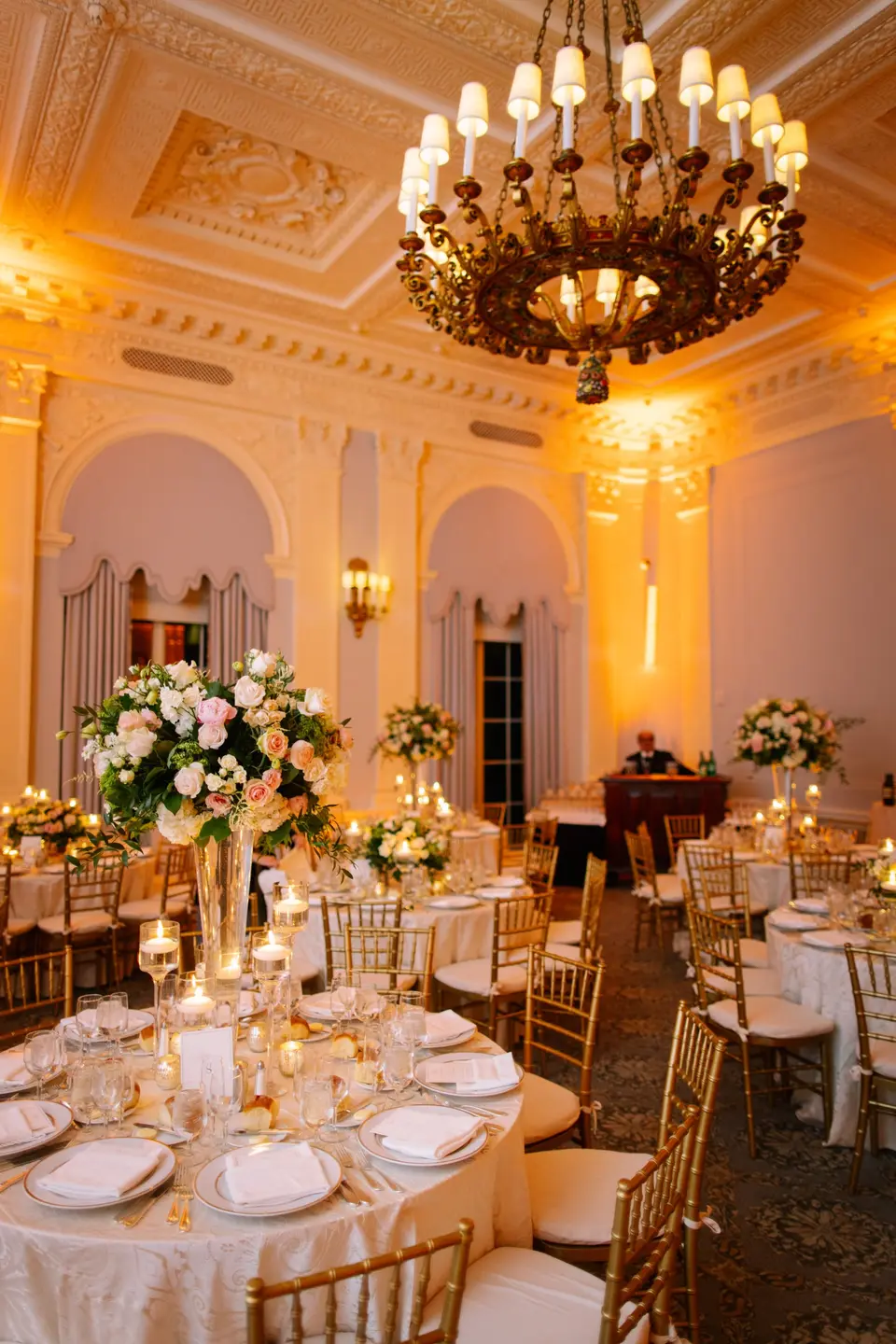 A round table set with candles and a tall arrangement of pink and white flowers beneath a bronze chandelier.