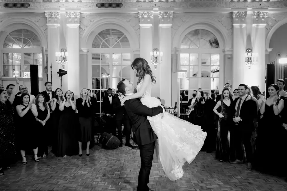 The groom lifts the bride on the dance floor as guests cheer. Black and white.