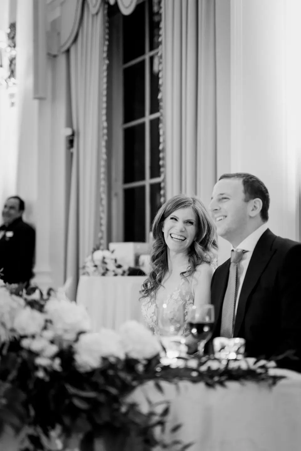 The bride and groom laugh at the sweetheart table during the toasts. Black and white.